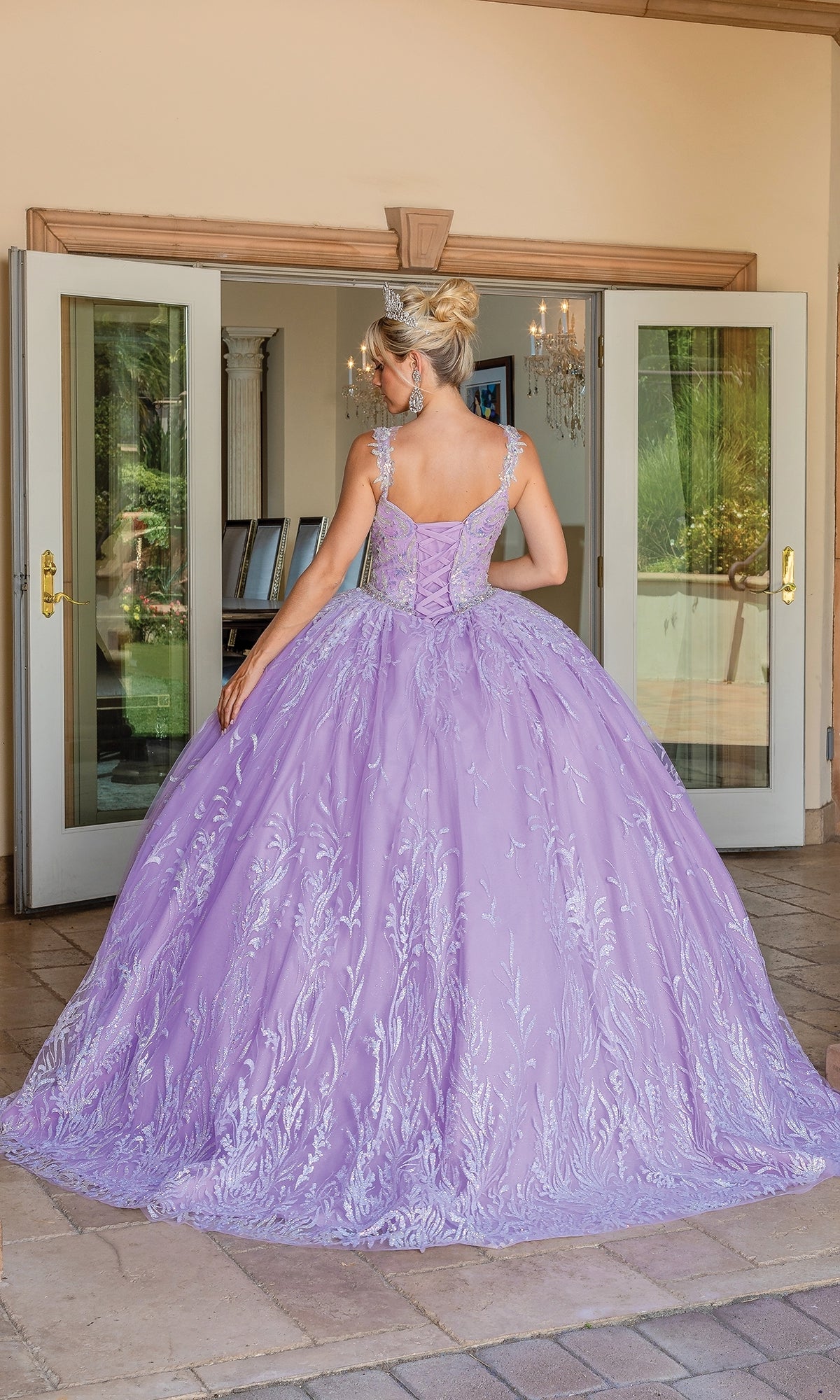 A woman faces away, wearing the Quinceanera Dress 1685 By Dancing Queen—an elegant lavender ball gown with embroidery and a sweetheart neckline—in front of open double doors to a brightly lit interior.